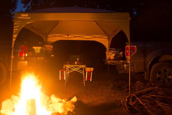 Camping setup. Burning campfire in the foreground. Two pickup trucks backed towards each other with tailgates down, acting as tables, and a camp table between them with a camping stove. Canopy centered between the tailgates.